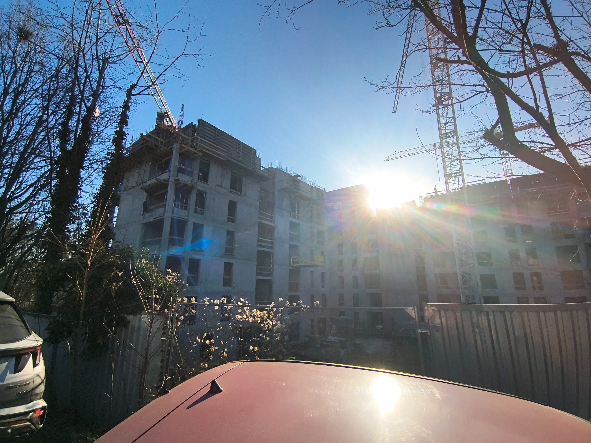 Wznoszący się budynek żelbetowy w trakcie budowy, widziany zza ogrodzenia i fragmentu samochodu, z żurawiami i jasnym słońcem w tle.