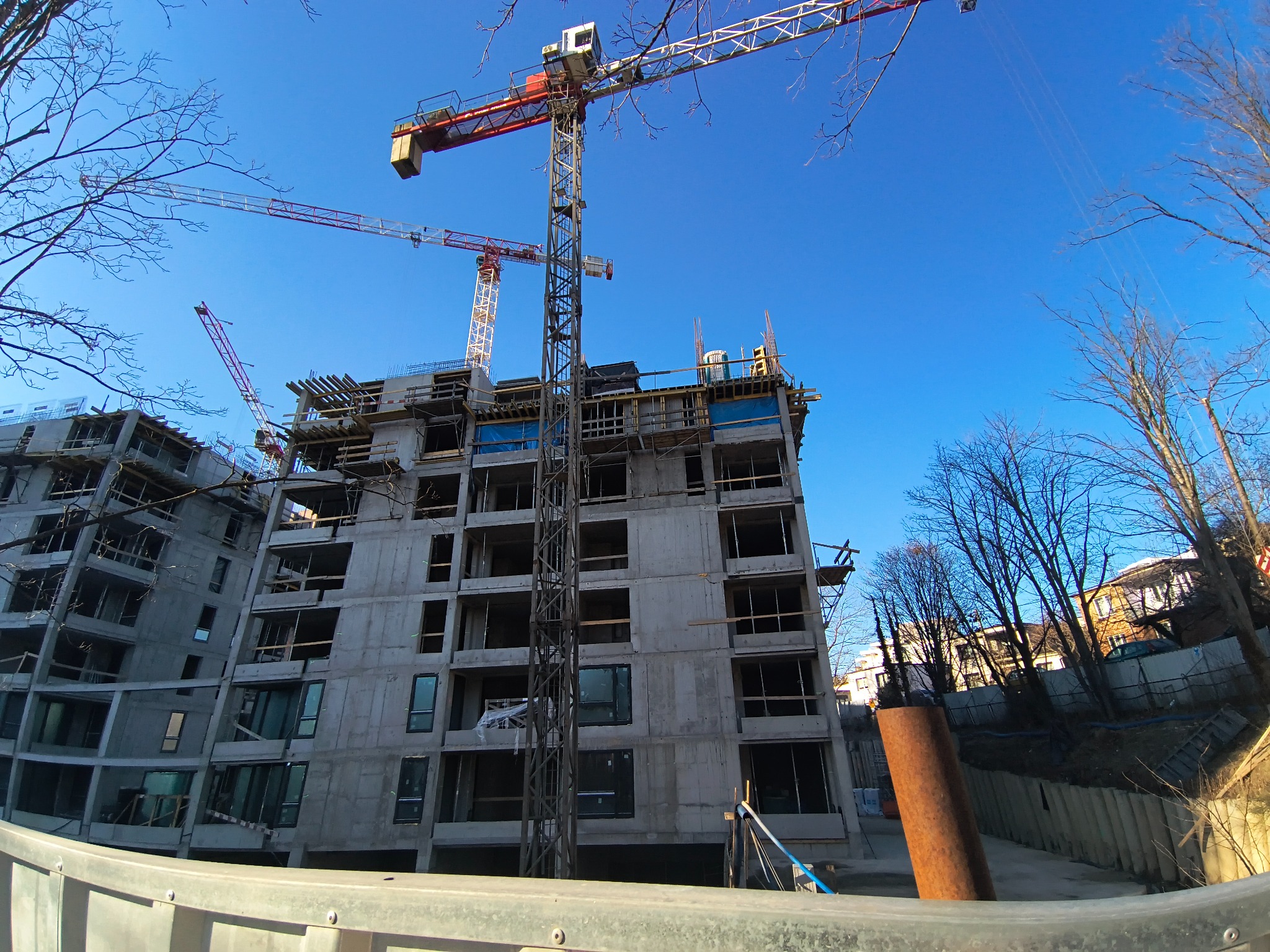 Wysoki budynek w stanie surowym, z widocznymi żelbetowymi konstrukcjami i dźwigami na tle błękitnego nieba. Widok z dołu, podkreślający skalę inwestycji.