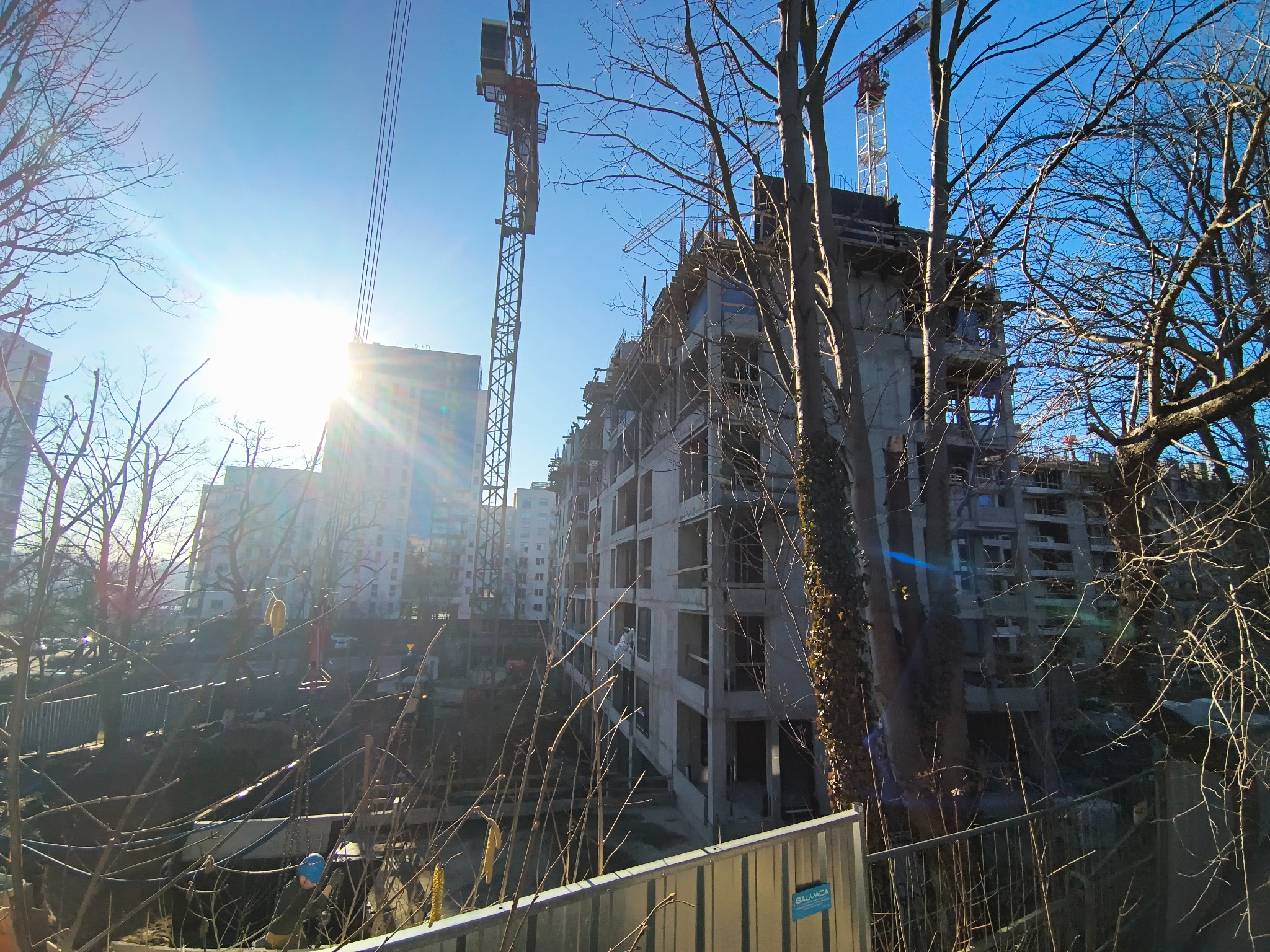 Wysoki budynek w trakcie budowy, widoczne rusztowania i żurawie. Surowa konstrukcja z betonu, w tle inne budynki i błękitne niebo. Ujęcie z dołu, zza ogrodzenia.
