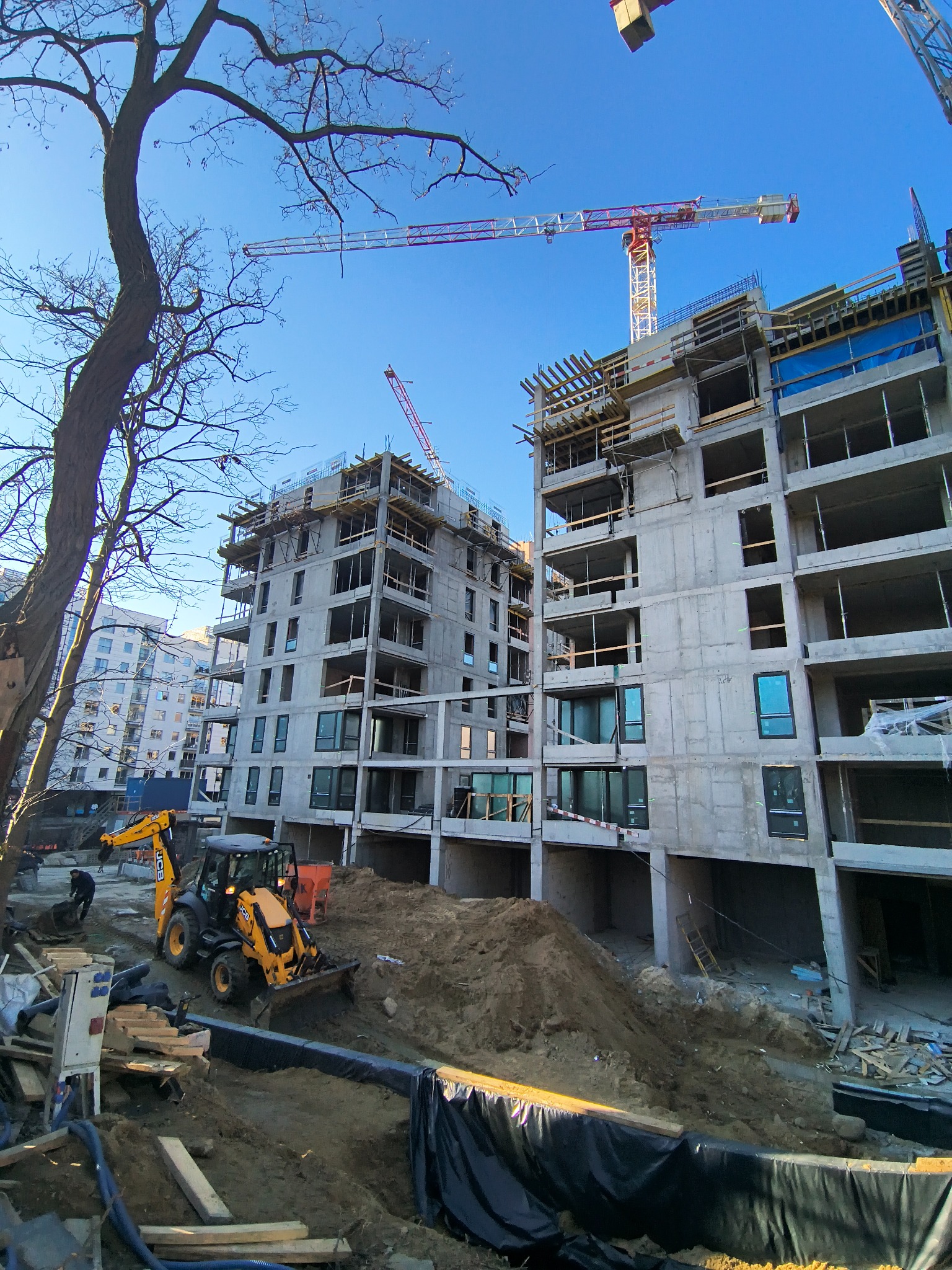 Budowa bloku z żelbetu w toku, widoczny dźwig i koparka JCB. Surowy stan, okna wstawione. Prace ziemne w toku. Błękitne niebo w tle.