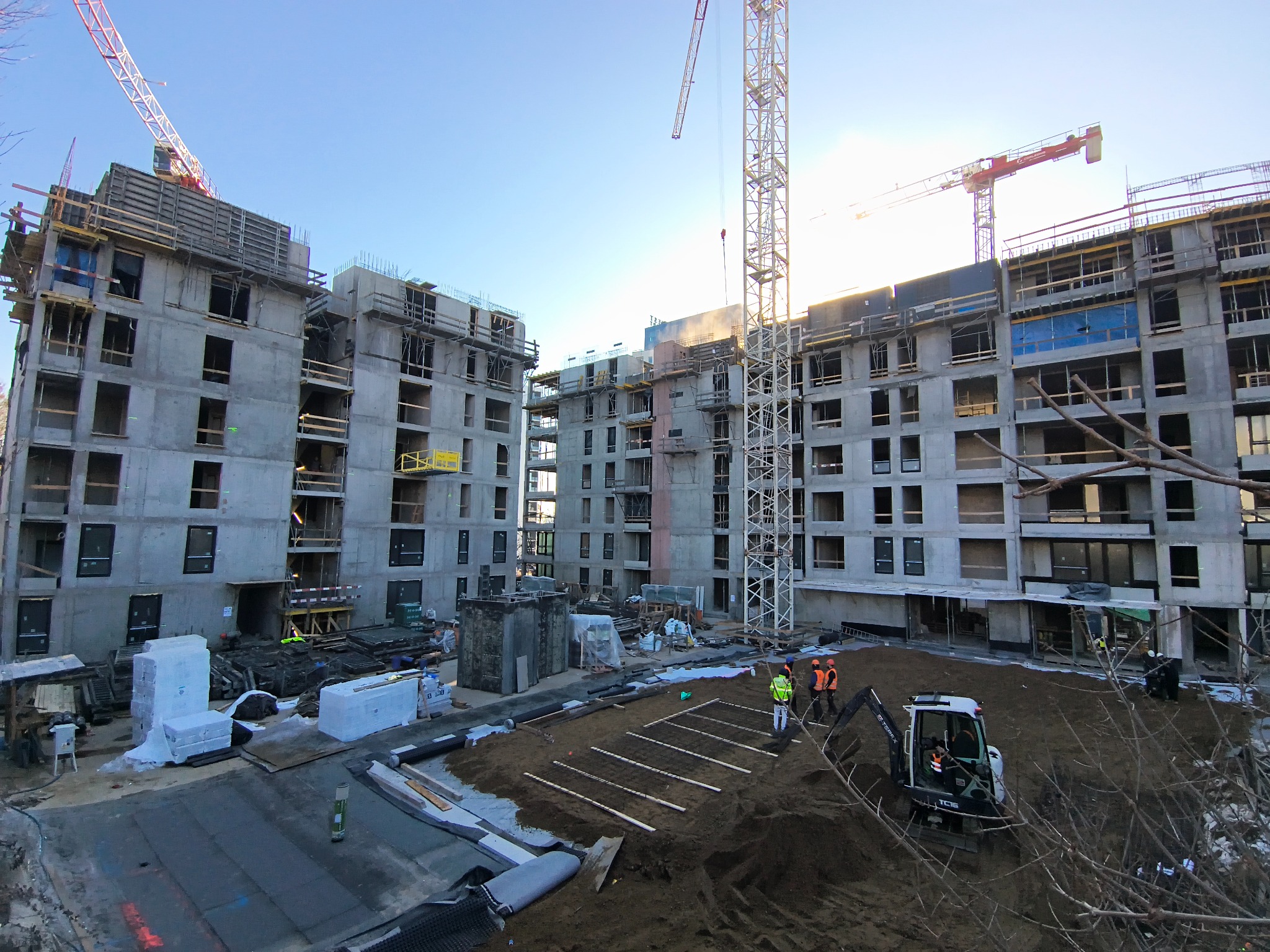 Wznoszenie surowych budynków wielorodzinnych z betonu, widoczne żurawie budowlane i ekipa robotników przygotowująca teren pod fundamenty.