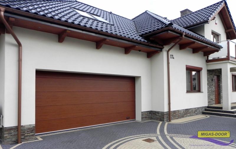 Nowoczesna brama garażowa segmentowa w kolorze drewna, zamontowana w białym budynku z ciemnym dachem i ozdobnym brukowanym podjazdem.