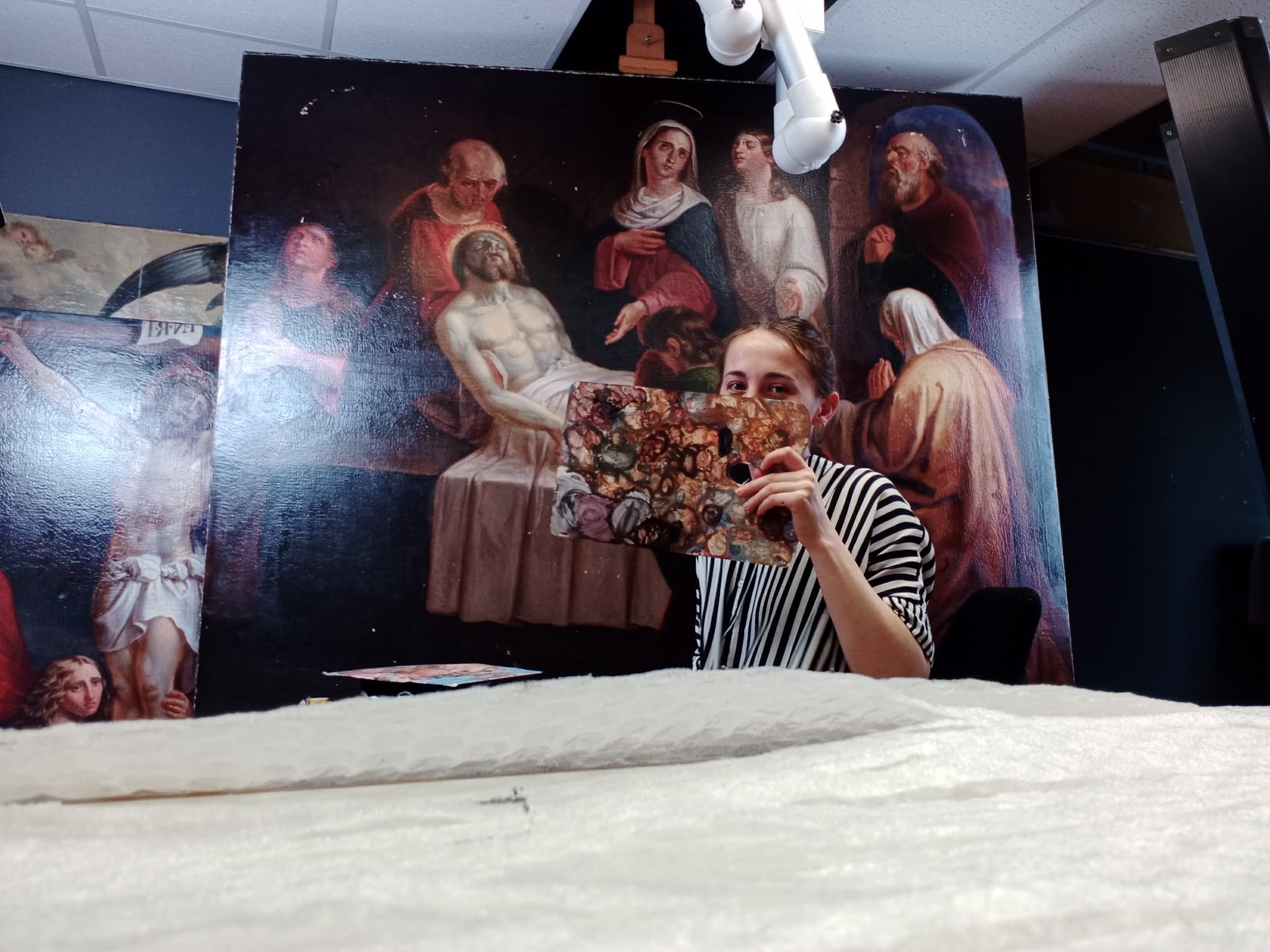 Kobieta w paski za paletą malarską, na tle wielkoformatowego obrazu olejnego z motywem religijnym. Widoczny fragment innego obrazu. Oświetlenie studyjne.