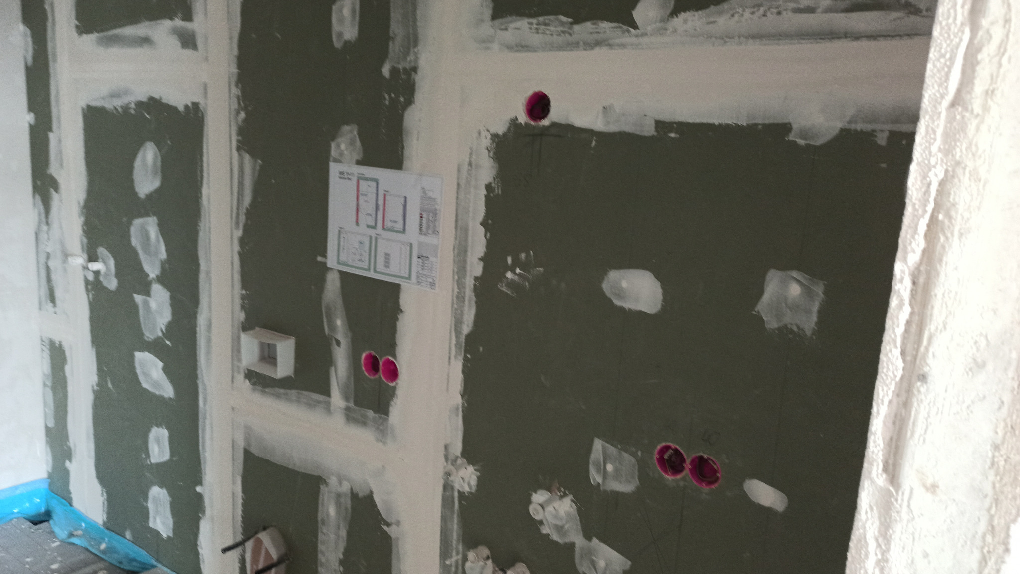 Ściana z płyt gipsowo-kartonowych w trakcie szpachlowania, widoczne otwory na gniazdka elektryczne i schemat instalacji na ścianie. Etap prac wykończeniowych.