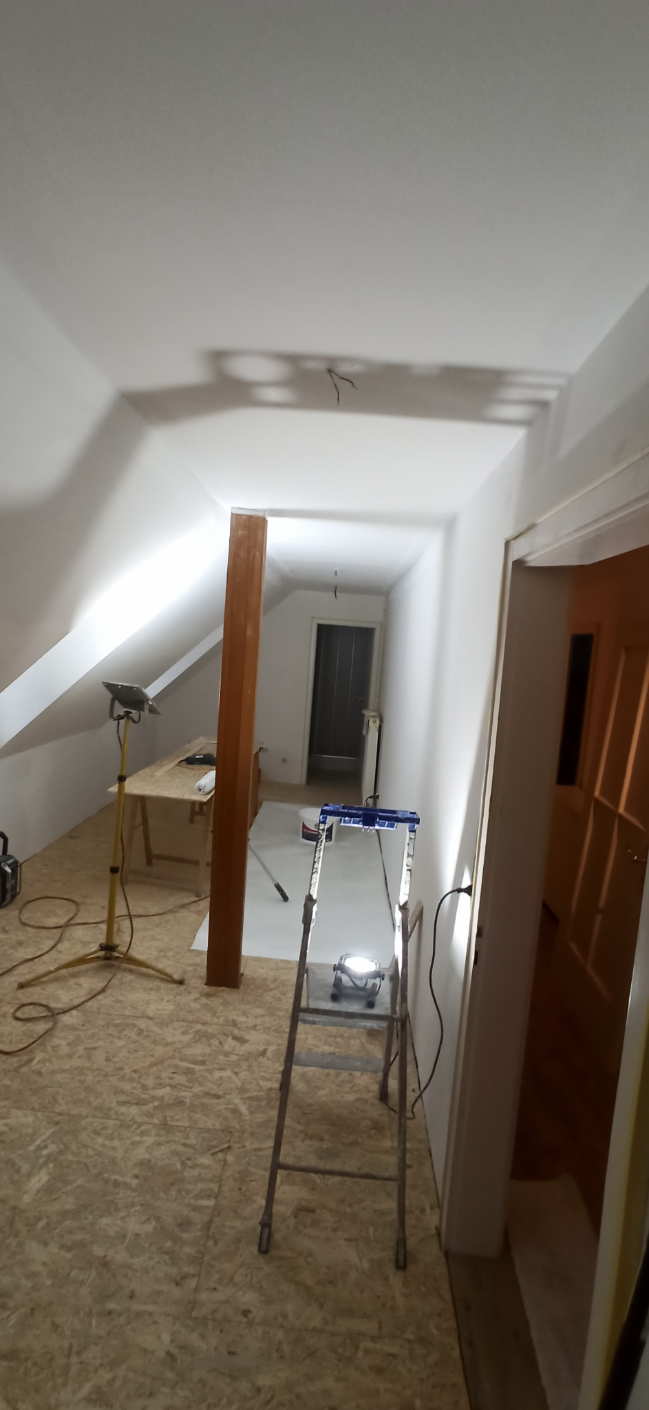 Poddasze w trakcie prac wykończeniowych: świeżo pomalowane ściany, surowe drewno na podłodze, drabina i lampa robocza. Widoczny fragment drzwi i drewniany słup.