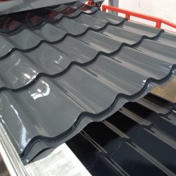 Transblach Jan Rusin - Błyszczące, szare dachówki ceramiczne w zbliżeniu, ułożone warstwami na metalowej konstrukcji. Widoczne detale falistego kształtu i gładkiej powierzchni.