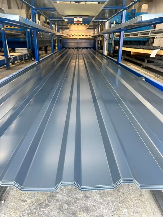 Szare arkusze blachy trapezowej ułożone na linii produkcyjnej, widok z perspektywy, z detalami profilowania i niebieską ramą maszyny w tle.