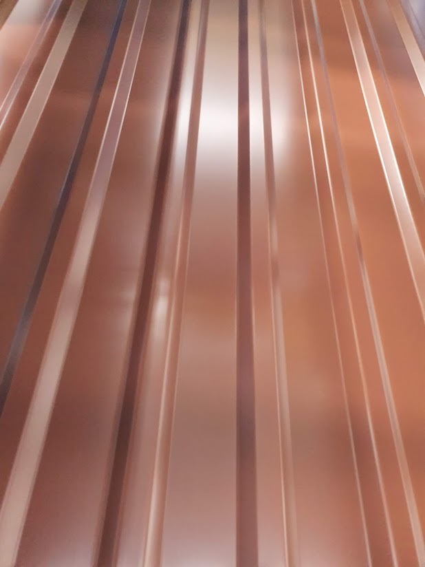 Błyszcząca, miedziana blacha trapezowa z wyraźnymi przetłoczeniami, sfotografowana pod kątem, ukazująca metaliczny połysk i regularny wzór. Nowoczesne pokrycie.