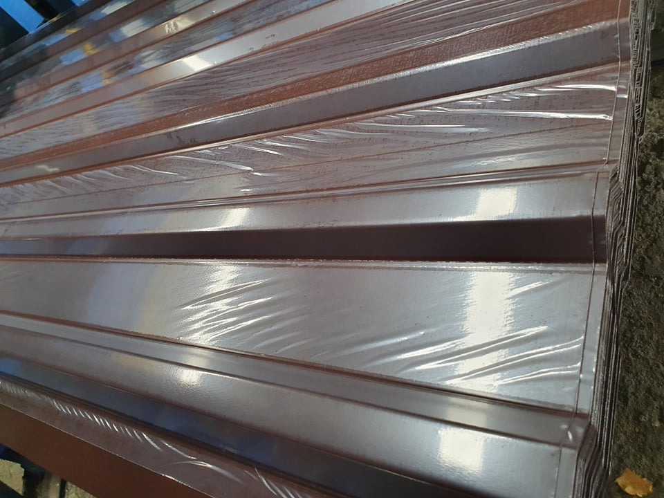 Stos brązowej blachy trapezowej zabezpieczonej folią, widoczny z boku, z delikatnym odbiciem światła. Elementy ułożone równolegle, tworząc powtarzalny wzór.