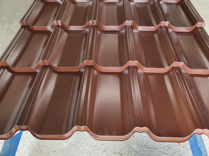 Błyszcząca, brązowa blacha dachowa w kształcie dachówki, ułożona na niebieskim podłożu. Widoczne regularne przetłoczenia i odbicia światła.