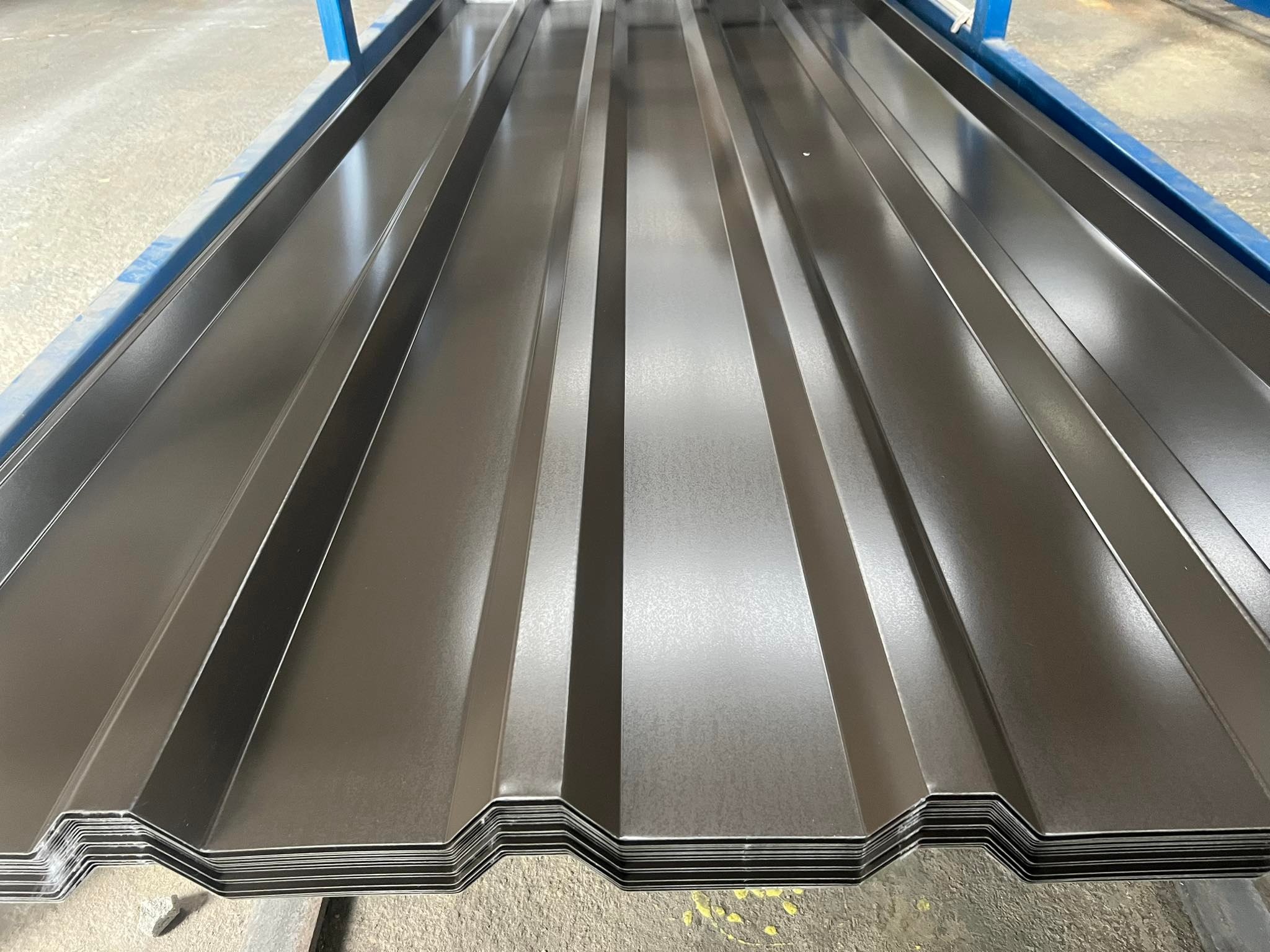 Stos arkuszy blachy trapezowej w kolorze grafitowym, ułożonych na niebieskiej konstrukcji. Widoczne charakterystyczne przetłoczenia i krawędzie arkuszy.