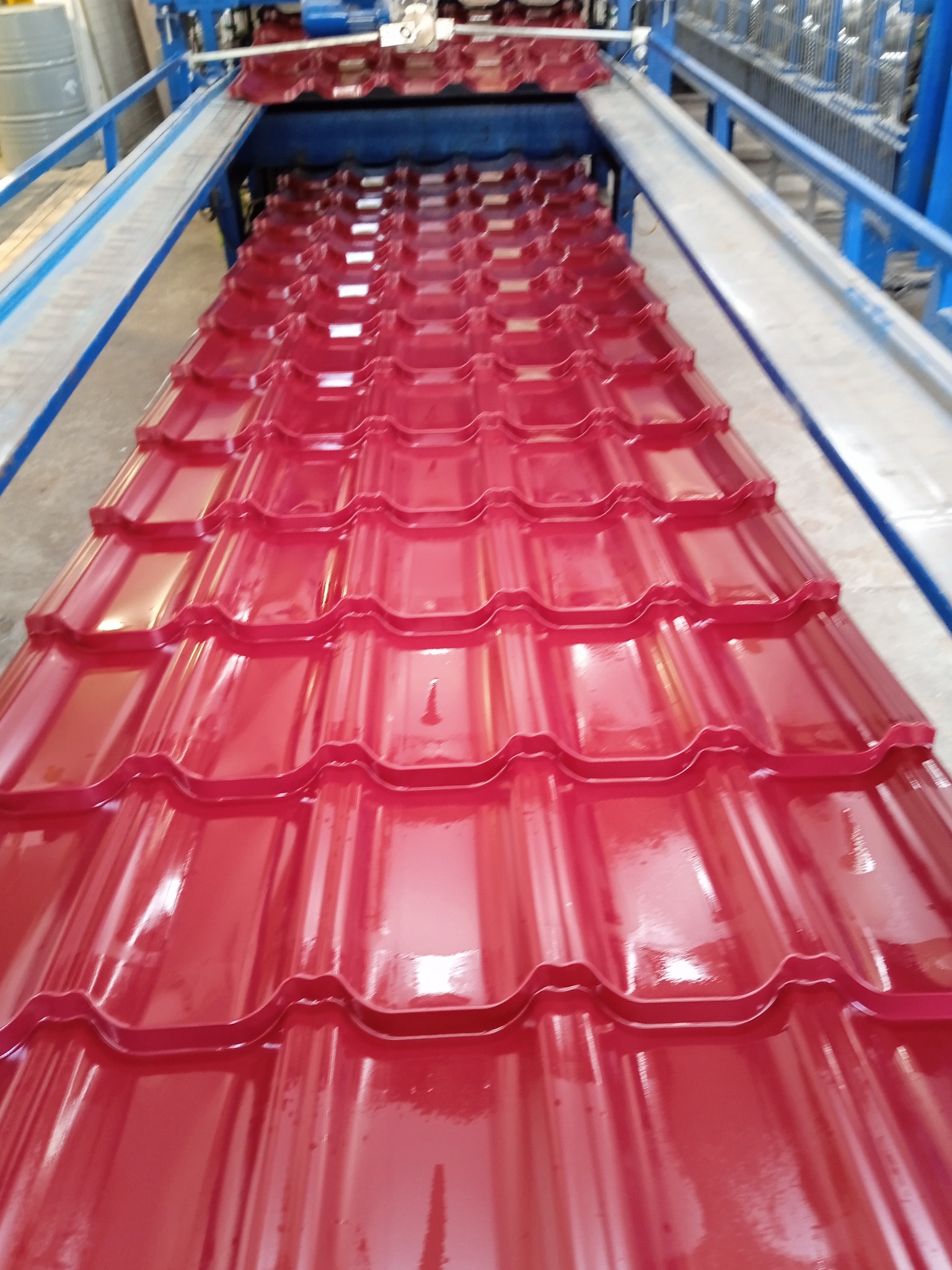 Czerwona blacha dachowa profilowana na maszynie, widok z góry. Błyszcząca powierzchnia, regularny wzór. Element linii produkcyjnej z niebieskimi elementami konstrukcyjnymi.
