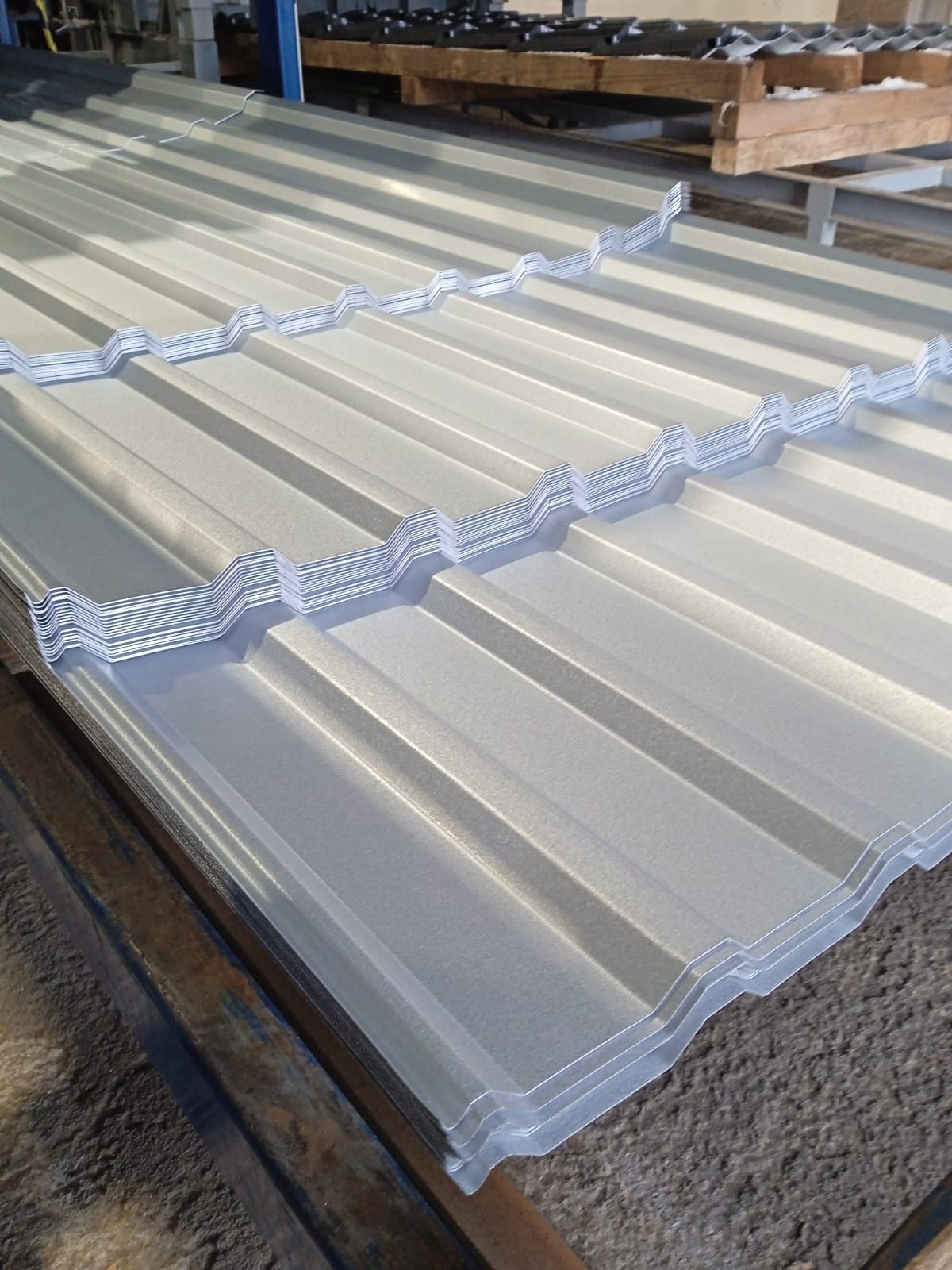 Stos srebrzystej blachy trapezowej z wyraźnym profilem falistym, ułożonej na metalowej konstrukcji. Widoczne detale faktury i odbicia światła na powierzchni.