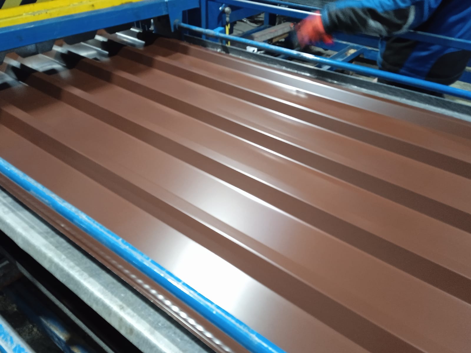Brązowa blacha trapezowa w procesie produkcji, widoczne elementy maszyny i dłoń pracownika w rękawicy. Ujęcie z bliska, ukazujące profilowanie arkusza.