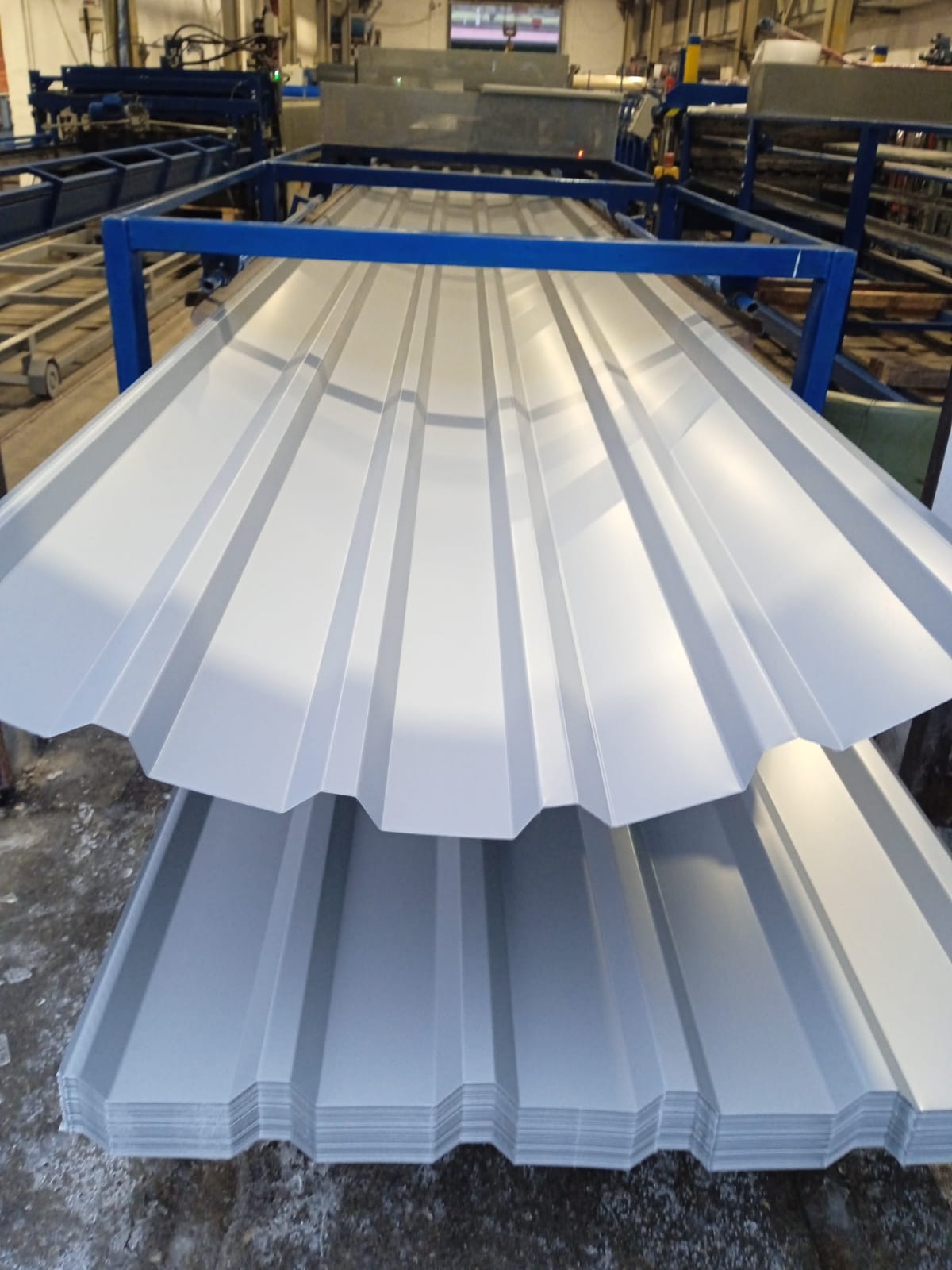 Stos blachy trapezowej w hali produkcyjnej, widoczne profile i niebieska konstrukcja maszyny w tle. Jasne oświetlenie podkreśla metaliczny połysk.
