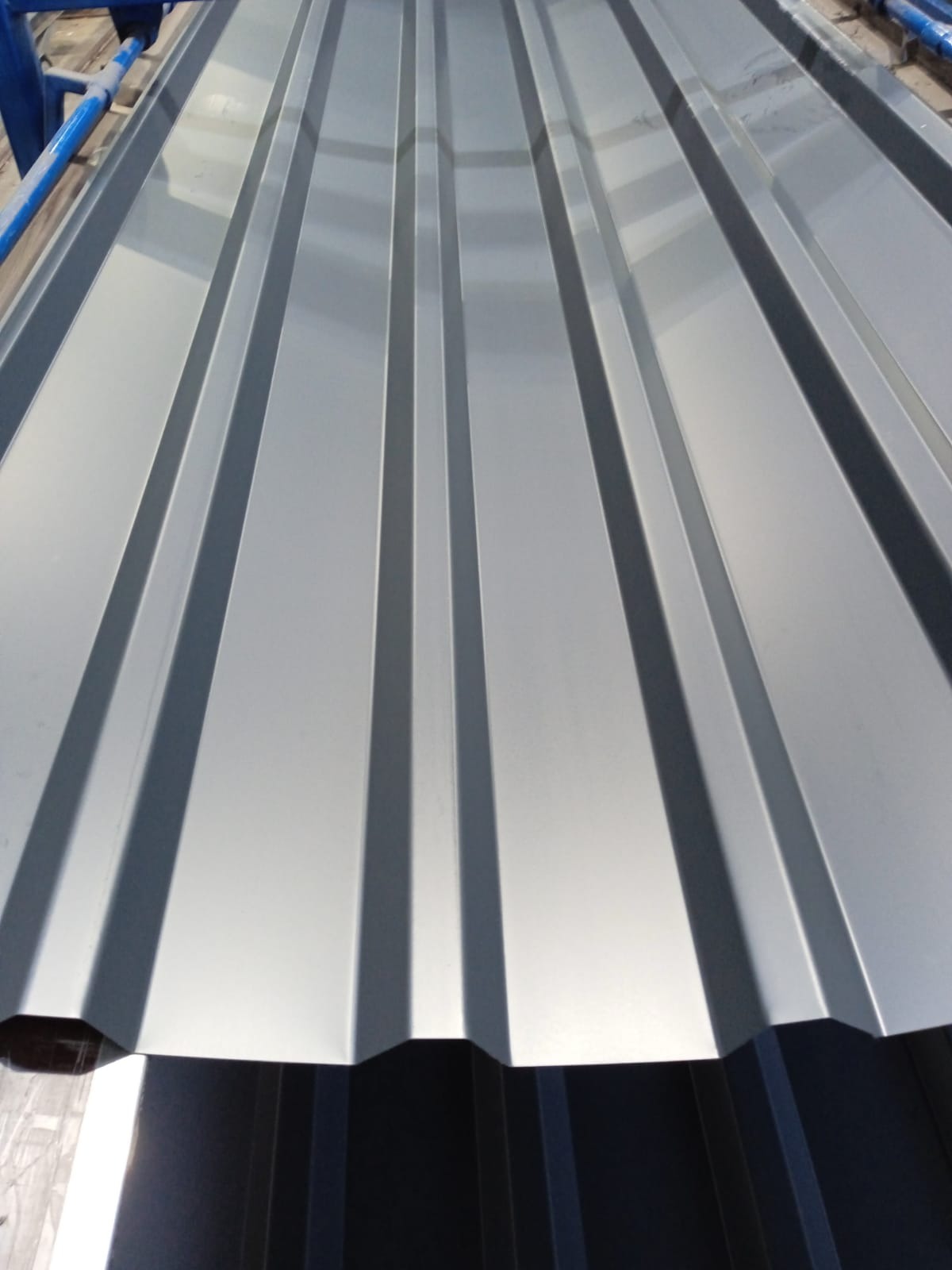 Błyszcząca, srebrna blacha trapezowa z wyraźnymi przetłoczeniami, ułożona na niebieskiej konstrukcji. Widoczne krawędzie arkuszy.