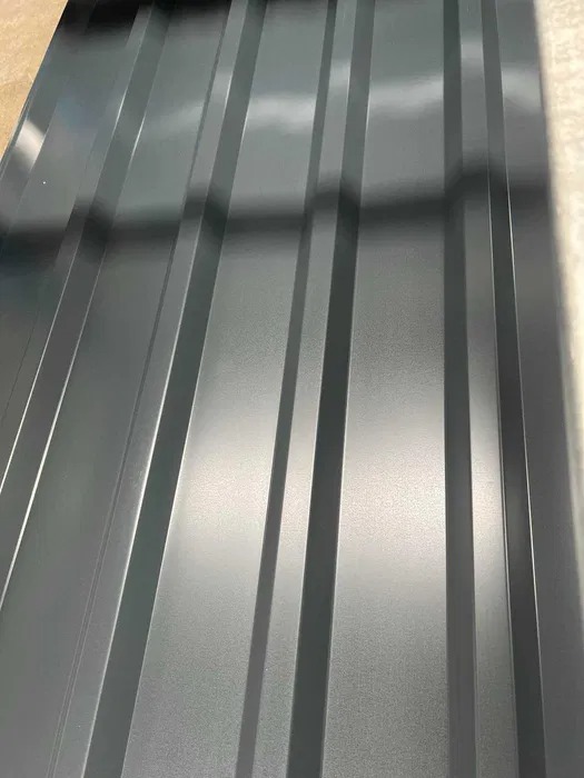 Szary arkusz blachy trapezowej z wyraźnym, regularnym wzorem przetłoczeń, leżący na jasnej powierzchni. Widoczne refleksy światła podkreślają metaliczną fakturę.