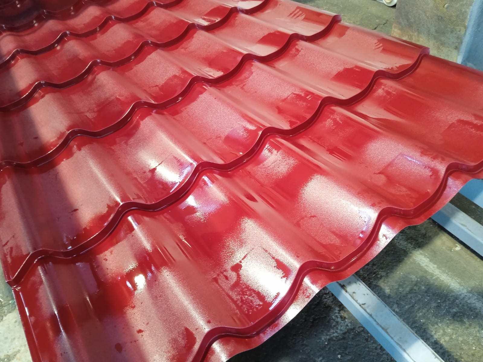 Błyszcząca, czerwona blacha dachowa o falistym profilu, ułożona na metalowej konstrukcji. Widoczne refleksy światła podkreślają intensywność koloru i fakturę materiału.