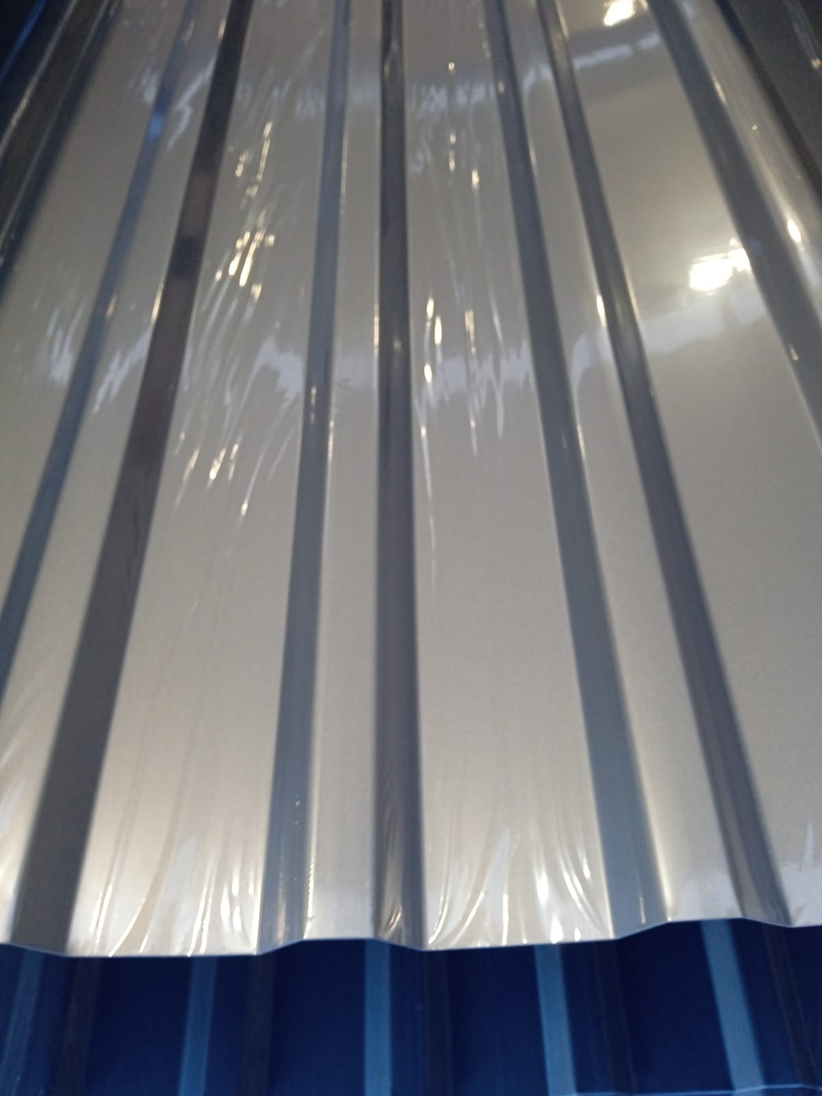 Błyszcząca, srebrna blacha trapezowa w folii ochronnej, widoczna z bliska pod kątem. Dolna krawędź w kolorze niebieskim. Oświetlenie studyjne.