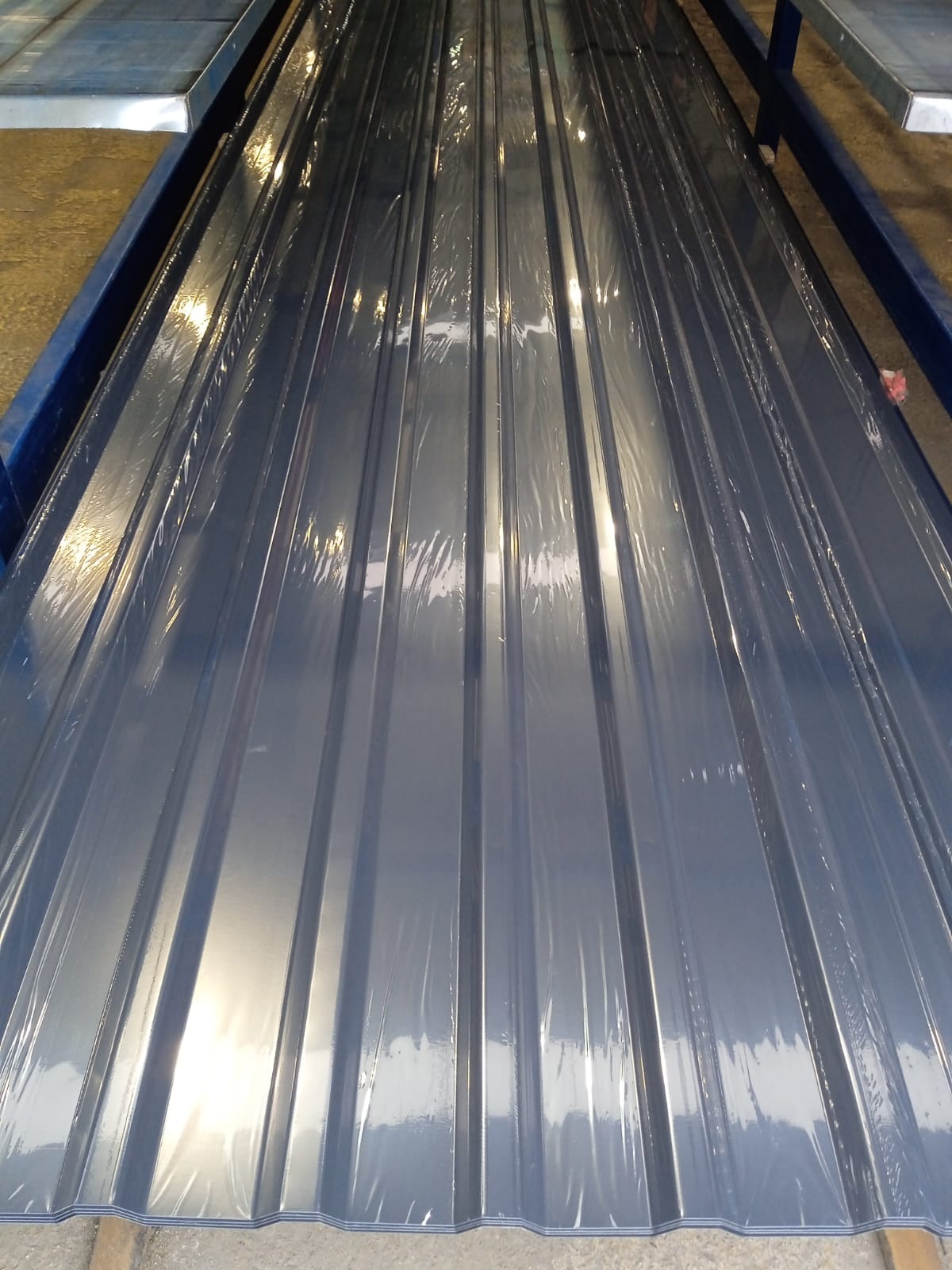 Szare arkusze blachy trapezowej, zabezpieczone folią, ułożone na metalowej konstrukcji. Widoczne regularne przetłoczenia i połyskująca powierzchnia.