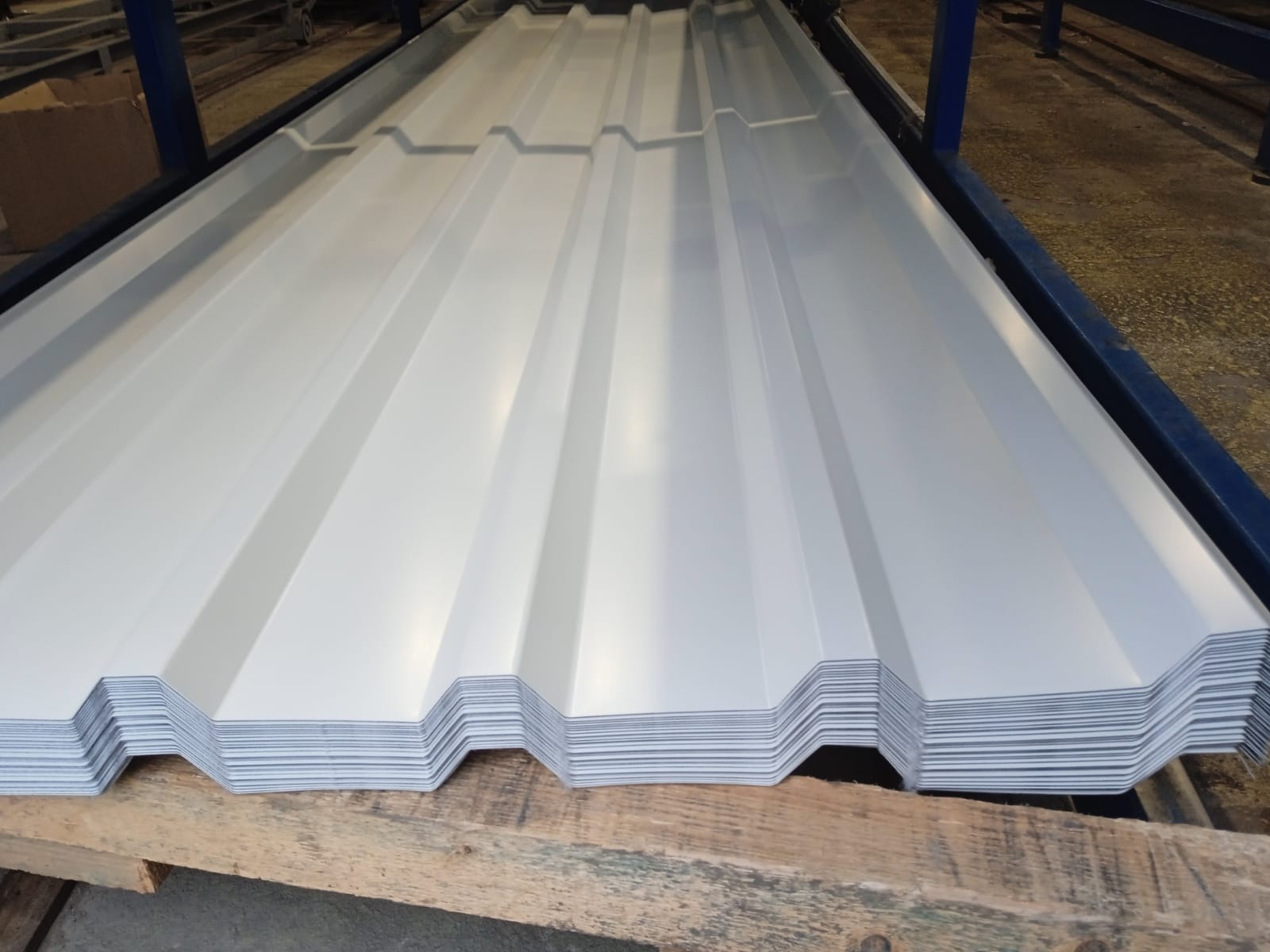 Stos białej blachy trapezowej ułożony na drewnianej palecie, widoczny z perspektywy. Blacha ma wyraźne, regularne przetłoczenia. W tle fragment konstrukcji stalowej.