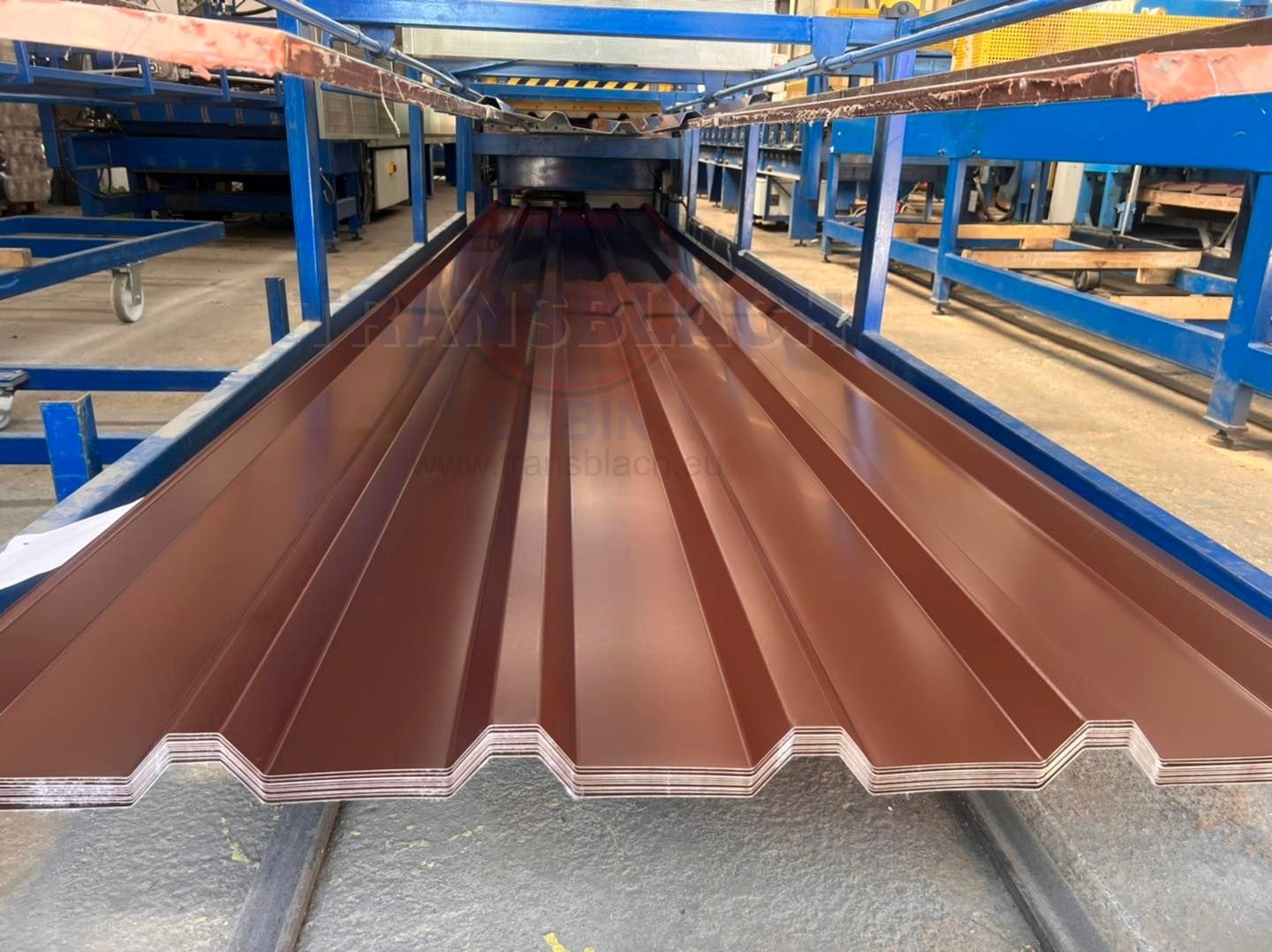 Brązowa blacha trapezowa na linii produkcyjnej, widoczne profilowanie i krawędzie. Metalowa konstrukcja w tle. Fabryczne oświetlenie.