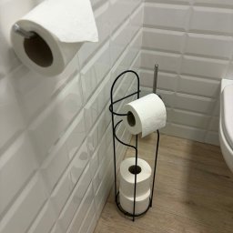 GlowClean Kinga Opach - Biała łazienka z podajnikiem na papier toaletowy. Na ścianie i podłodze jasne płytki. Widoczny fragment toalety. Dbałość o detale w czystym biurze.