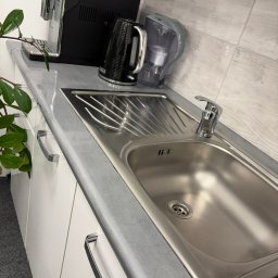 GlowClean Kinga Opach - Błyszczący, stalowy zlew wkomponowany w szary blat kuchenny, uzupełniony o ekspres do kawy, czajnik i filtr do wody. Minimalistyczny design, jasne płytki na ścianie.