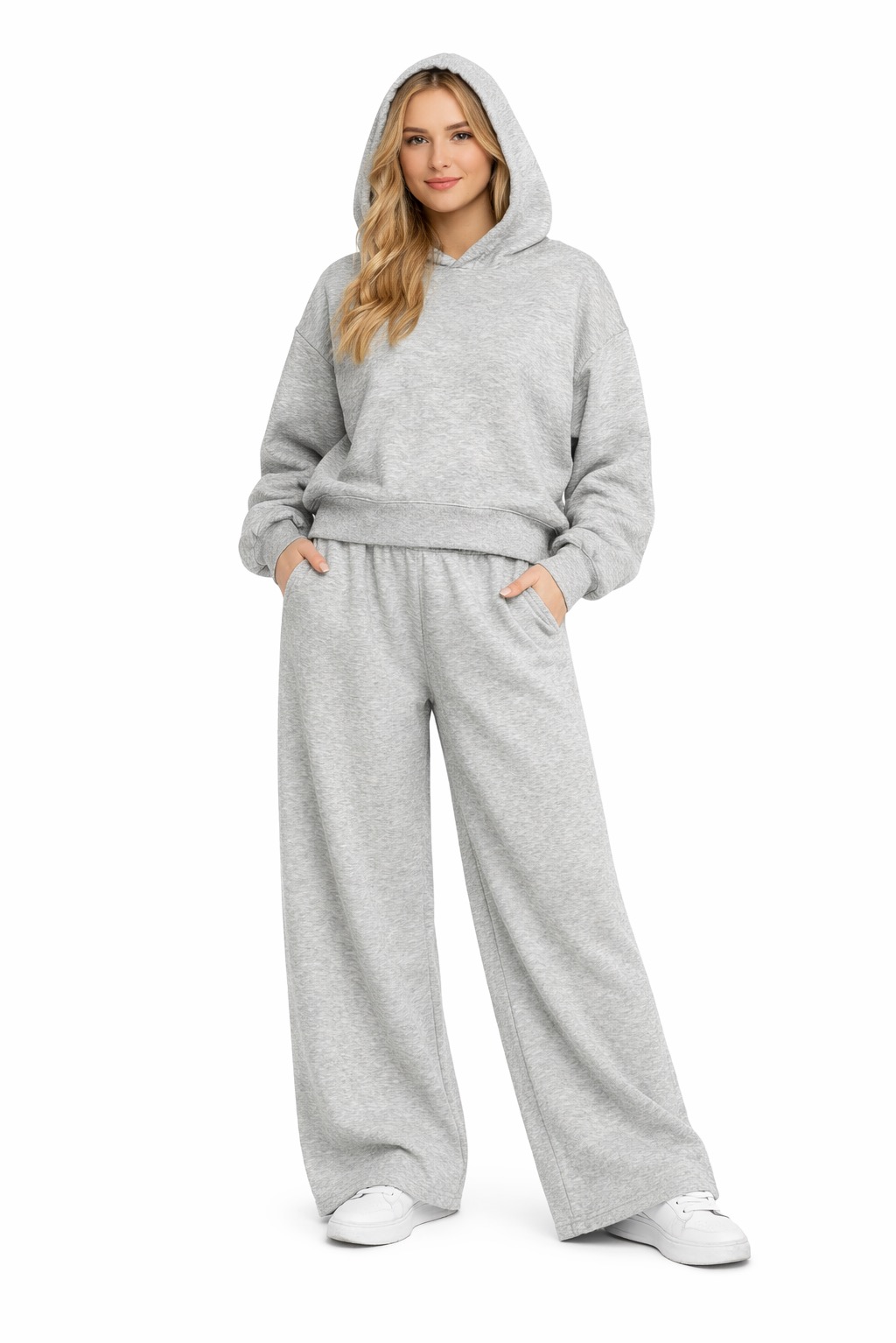 Kobieta w szarym dresie z kapturem i szerokimi spodniami, ręce w kieszeniach, na białym tle. Casualowy styl, widoczne detale materiału i szwy.