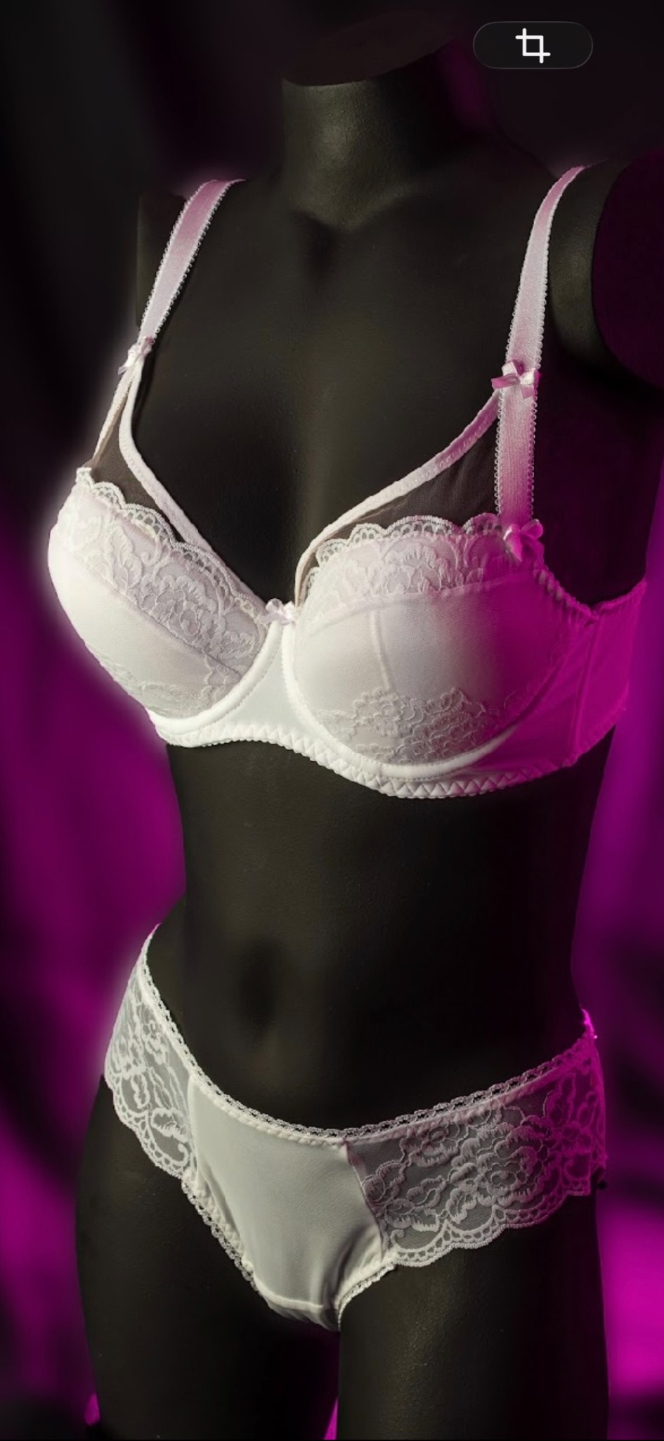 Biały komplet bielizny z koronką, składający się z biustonosza i majtek, prezentowany na czarnym manekinie na tle fuksjowego światła.