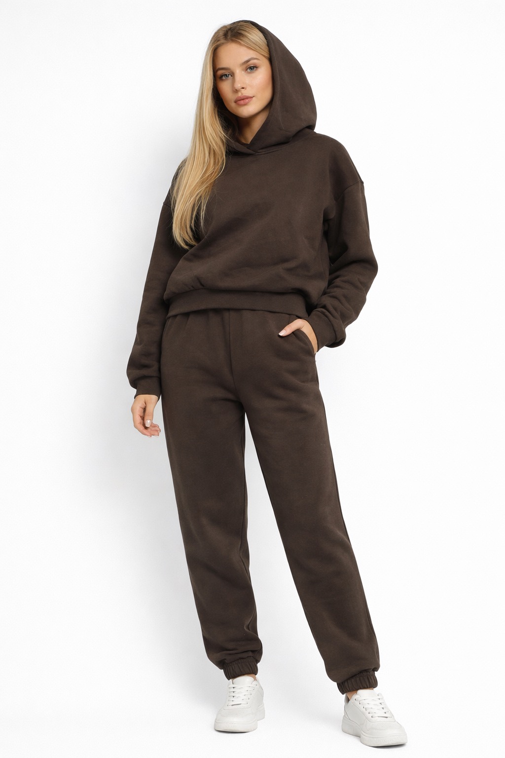 Modelka prezentuje brązowy, dresowy komplet z kapturem na białym tle. Swobodny krój, dresowe spodnie ze ściągaczami i białe sneakersy. Casualowy styl.