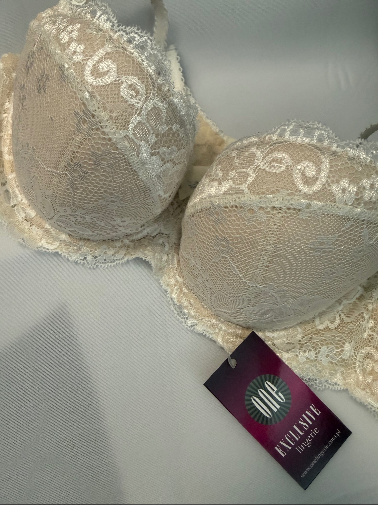 Beżowy biustonosz z koronkowymi zdobieniami, leżący na białym tle, z widoczną metką marki 'ONE Exclusive lingerie'. Delikatne światło podkreśla detale.