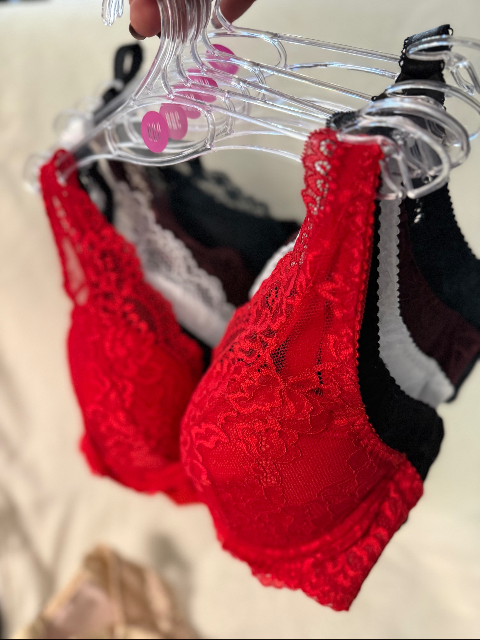 Koronkowe biustonosze w kolorach czerwonym, białym i czarnym, wiszące na przezroczystych wieszakach. Widoczne metki z rozmiarami. Delikatna faktura materiału.