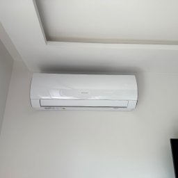 Biała klimatyzacja Sinclair na ścianie w jasnym wnętrzu. Widoczny fragment sufitu z oświetleniem i czarny telewizor w rogu kadru. Minimalistyczny design.