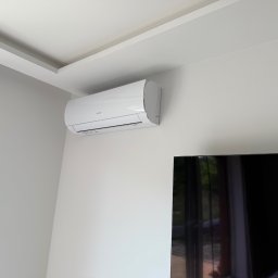 Biały klimatyzator ścienny zamontowany w rogu pokoju, widoczny fragment sufitu z dekoracyjną listwą i fragment czarnego telewizora. Minimalistyczny design wnętrza.