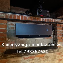 Czarny klimatyzator ścienny z wyświetlaczem temperatury (16°C) zamontowany na ceglanej ścianie w pomieszczeniu z widocznym projektorem sufitowym.