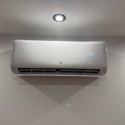 Biały klimatyzator AUX z wyświetlaczem temperatury (22°C) zamontowany na jasnej ścianie pod okrągłym oświetleniem sufitowym. Minimalistyczny design.