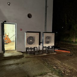 SolidMark Marek Papis - Dwa zainstalowane, białe moduły pompy ciepła Toshiba na zewnątrz budynku, widoczne rury i otwarte drzwi do pomieszczenia z instalacjami. Nocne ujęcie.