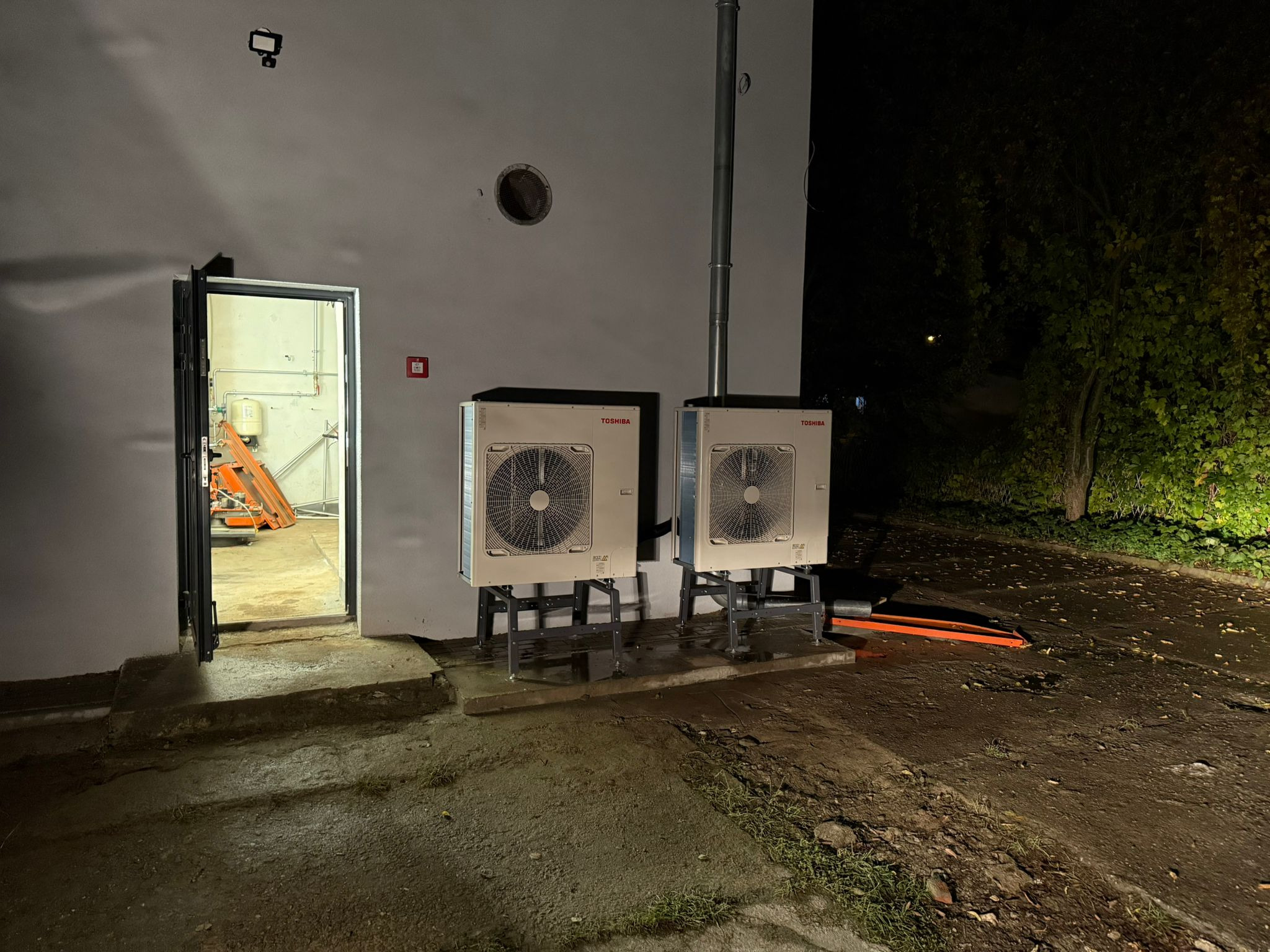 Dwa zainstalowane, białe moduły pompy ciepła Toshiba na zewnątrz budynku, widoczne rury i otwarte drzwi do pomieszczenia z instalacjami. Nocne ujęcie.