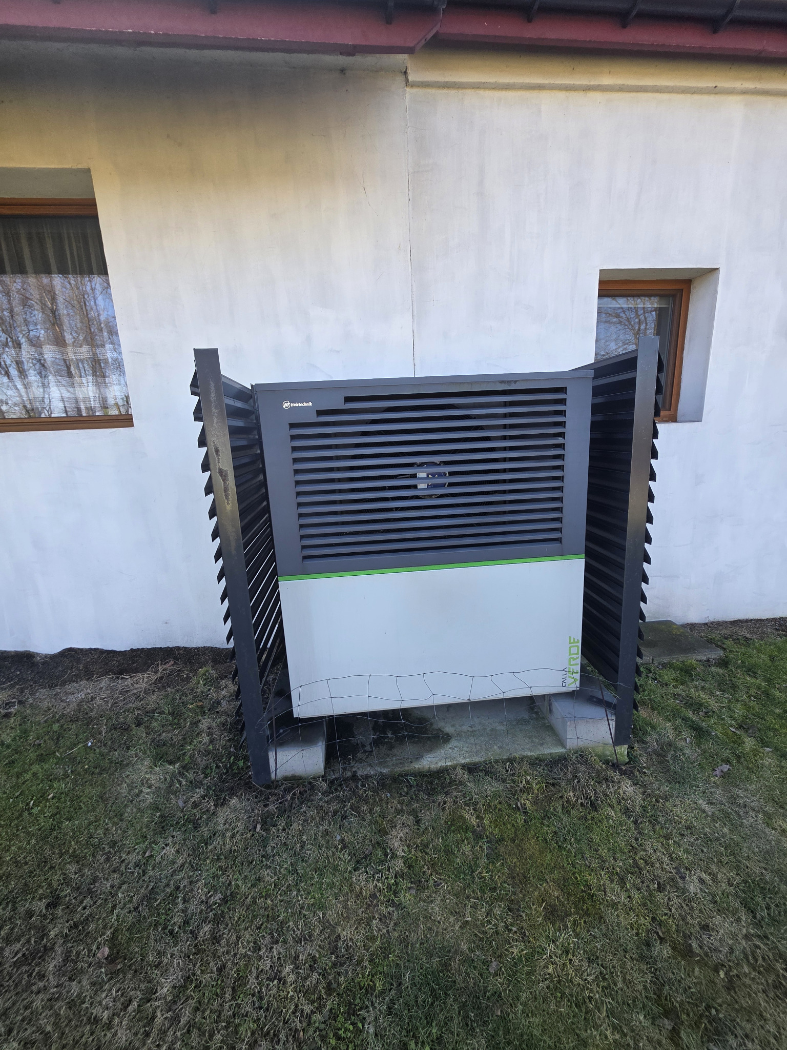 Pompa ciepła Holztechnik na zewnątrz budynku, zabezpieczona metalową osłoną. Widoczne okna budynku w tle. Ujęcie z poziomu gruntu.