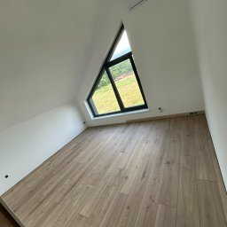 Ihor Dmytriv - Jasne panele podłogowe w pokoju na poddaszu z trójkątnym oknem i klimatyzacją. Ściany pomalowane na biało, widok na zewnątrz na zielone pole. Minimalistyczny design.