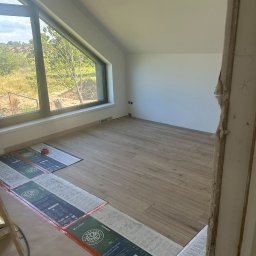 Ihor Dmytriv - Jasne wnętrze poddasza z ułożoną drewnopodobną podłogą, duże okno z widokiem na zieleń. Częściowo widoczne panele podłogowe w opakowaniach. Ściany pomalowane na biało.