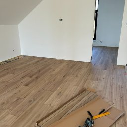 Ihor Dmytriv - Jasne panele podłogowe ułożone w pokoju na poddaszu, widoczne narzędzia pracy: wiertarka, młotek, kątownik. Ściany pomalowane na biało, widoczne puszki elektryczne.