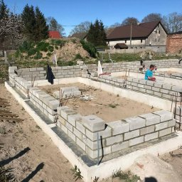 Ihor Dmytriv - Wznoszenie fundamentu z bloczków betonowych na piasku, widoczne zbrojenia i pracownik w tle. Budowa domu w słoneczny dzień.