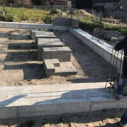 Ihor Dmytriv - Wznoszenie fundamentu z bloczków betonowych na piaszczystym podłożu. Widoczne zbrojenia i częściowo ukończona ściana fundamentowa. W tle dom z czerwoną dachówką.