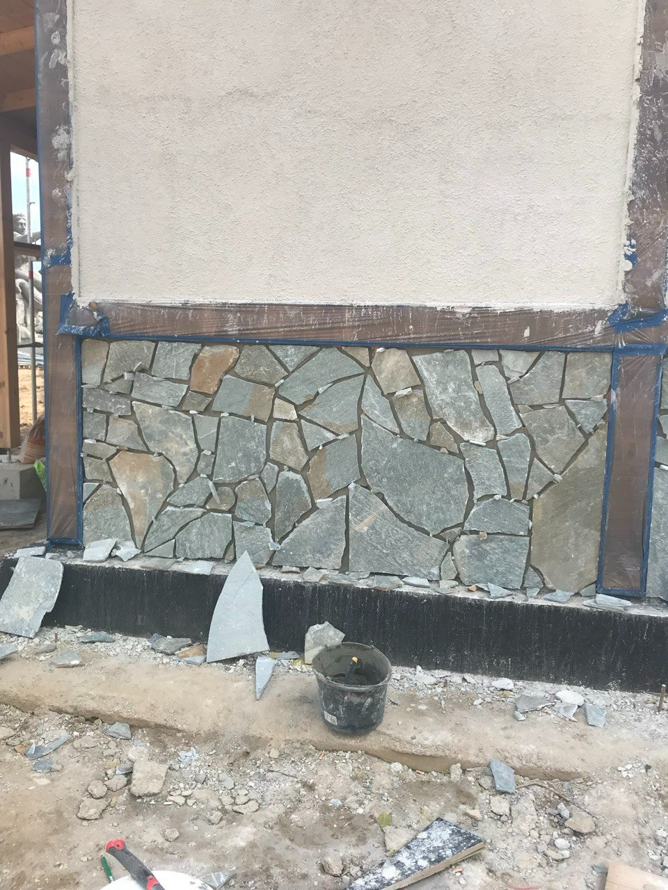 Wykończenie ściany zewnętrznej nieregularnym kamieniem elewacyjnym. Widoczne fragmenty niezabudowane, wiadro z zaprawą i narzędzia. Prace w toku.