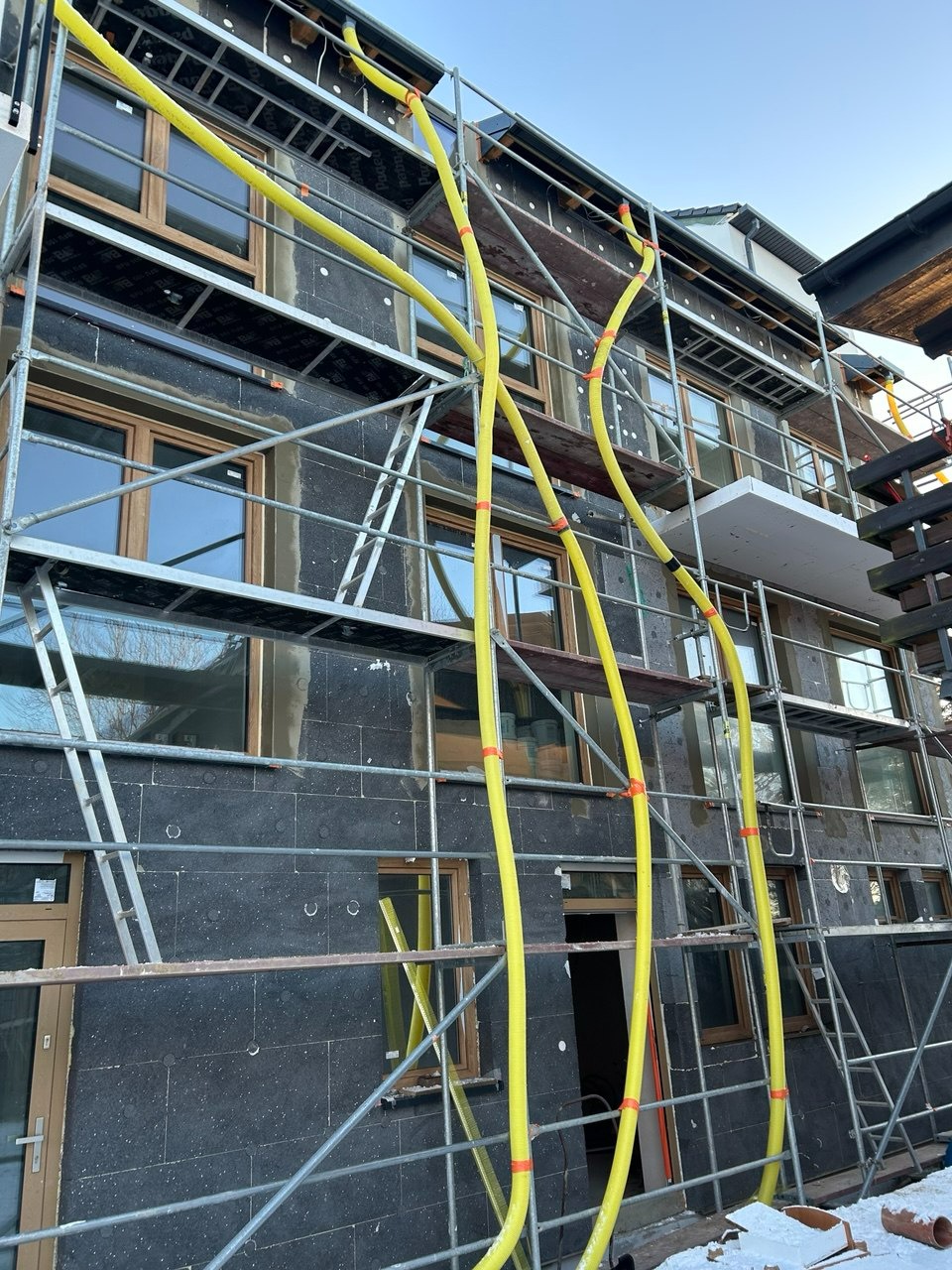 Ocieplanie elewacji budynku wielorodzinnego z użyciem rusztowań i żółtych węży wentylacyjnych. Widoczne płyty izolacyjne na ścianach.