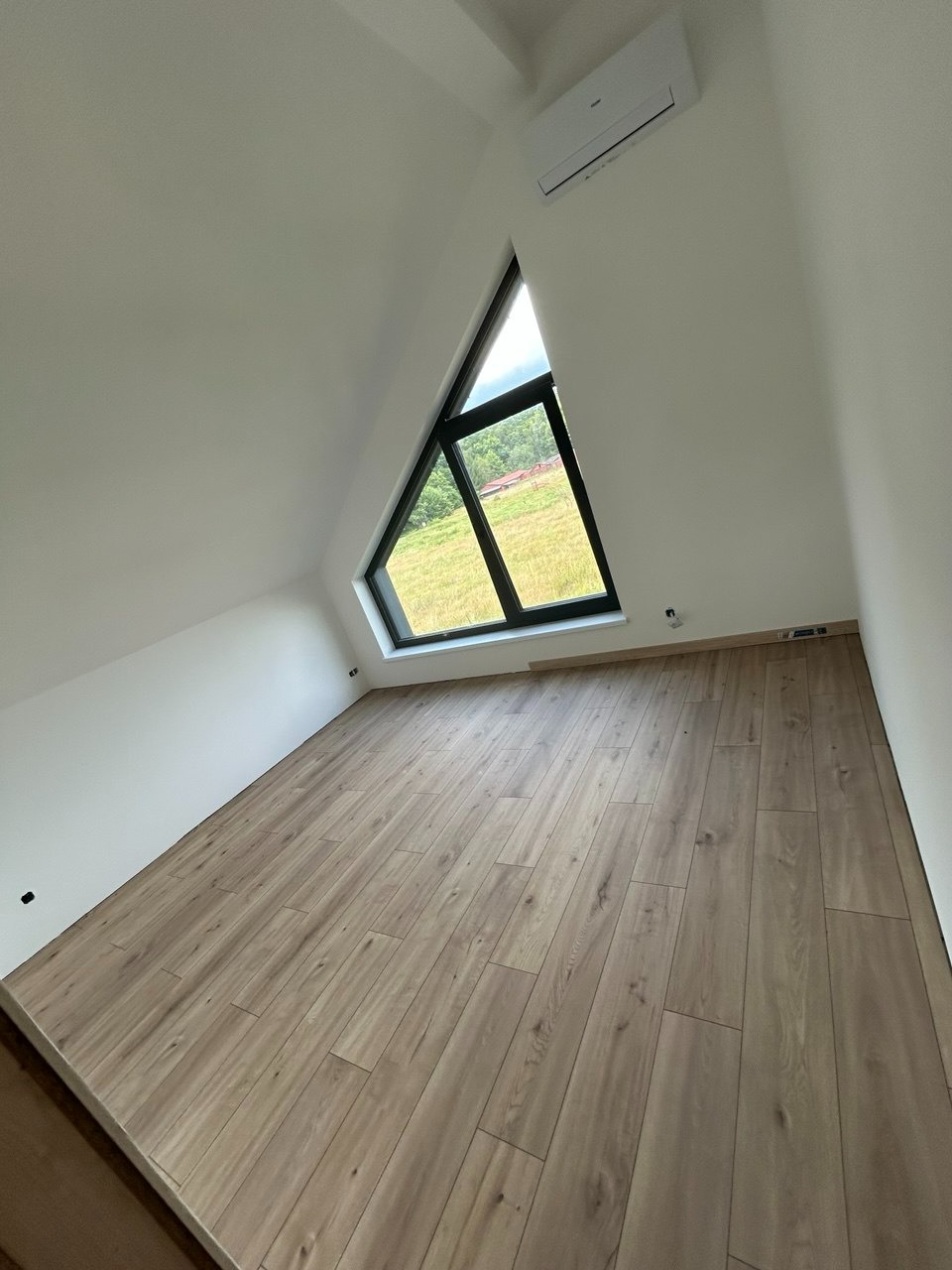 Jasne panele podłogowe w pokoju na poddaszu z trójkątnym oknem i klimatyzacją. Ściany pomalowane na biało, widok na zewnątrz na zielone pole. Minimalistyczny design.