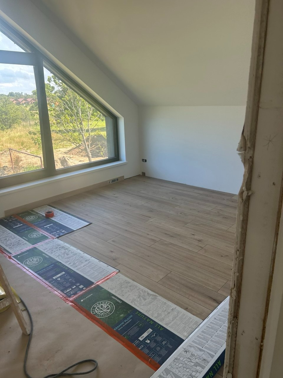 Jasne wnętrze poddasza z ułożoną drewnopodobną podłogą, duże okno z widokiem na zieleń. Częściowo widoczne panele podłogowe w opakowaniach. Ściany pomalowane na biało.