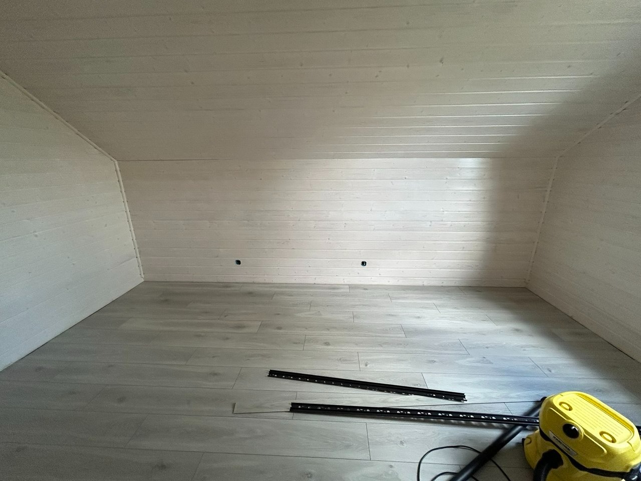 Wykończone poddasze z boazerią i panelami podłogowymi w odcieniach bieli i szarości. Widoczne listwy przypodłogowe i odkurzacz. Ukończony etap prac.